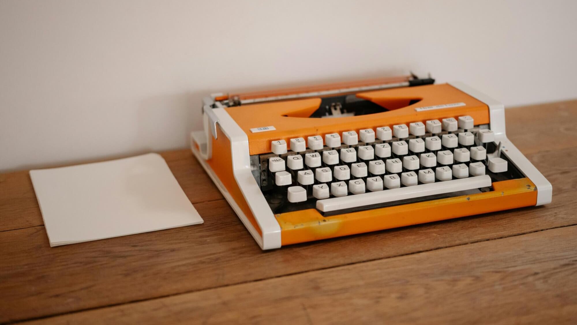 Orangefarbene Schreibmaschine auf einem Holztisch neben einem leeren Blatt Papier als Symbol für Texten mit KI.